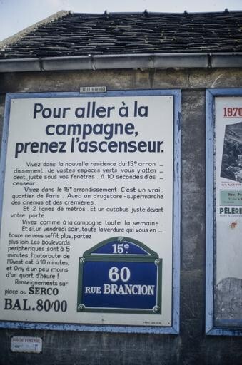 Affiche publicitaire sur un mur, promouvant une nouvelle résidence 60 rue Brancion à Paris. L’affiche porte l’image d’une plaque de rue parisienne et le long texte suivant : « Pour aller à la campagne, prenez l’ascenseur. Vivez dans la nouvelle résidence du 15e arrondissement : de vastes espaces verts vous y attendent, juste sous vos fenêtres. À 10 secondes d’ascenseur. Vivez dans le 15e arrondissement. C’est un vrai quartier de Paris. Avec un drugstore-supermarché, des cinémas et des crémières. Et 2 lignes de métro. Et un autobus juste devant votre porte. Vivez comme à la campagne toute la semaine. Et si, un vendredi soir, toute la verdure qui vous entoure ne vous suffit plus, partez plus loin. Les boulevards périphériques sont à 5 minutes, l’autoroute de l’Ouest est à 10 minutes, et Orly à un peu moins d’un quart d’heure ! Renseignements sur place ou SERCO. BAL.80/00 »